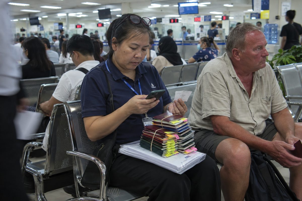 A visa agent at Chaeng Wattana immigration with a stack of client passports and a queue ticket, a foreign client waiting beside her.